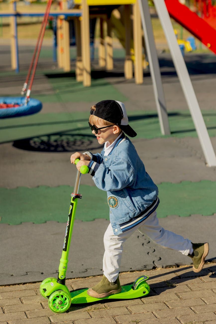 A playful child rides a green scooter outdoors on a sunny day, wearing a denim jacket and cap.